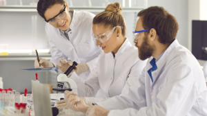 Building brilliant culture in Biotech: image depicts 3 scientists wearing lab coats and safety goggles working together in a lab space. One lady looks through a microscope, with another lady is taking notes on a clipboard.