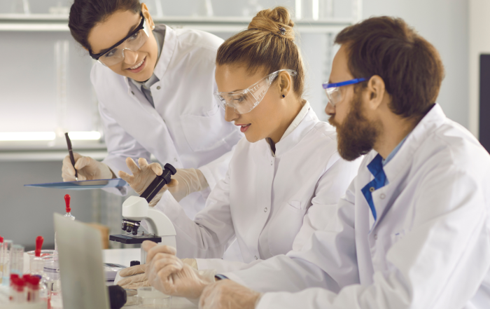 Building brilliant culture in Biotech: image depicts 3 scientists wearing lab coats and safety goggles working together in a lab space. One lady looks through a microscope, with another lady is taking notes on a clipboard.