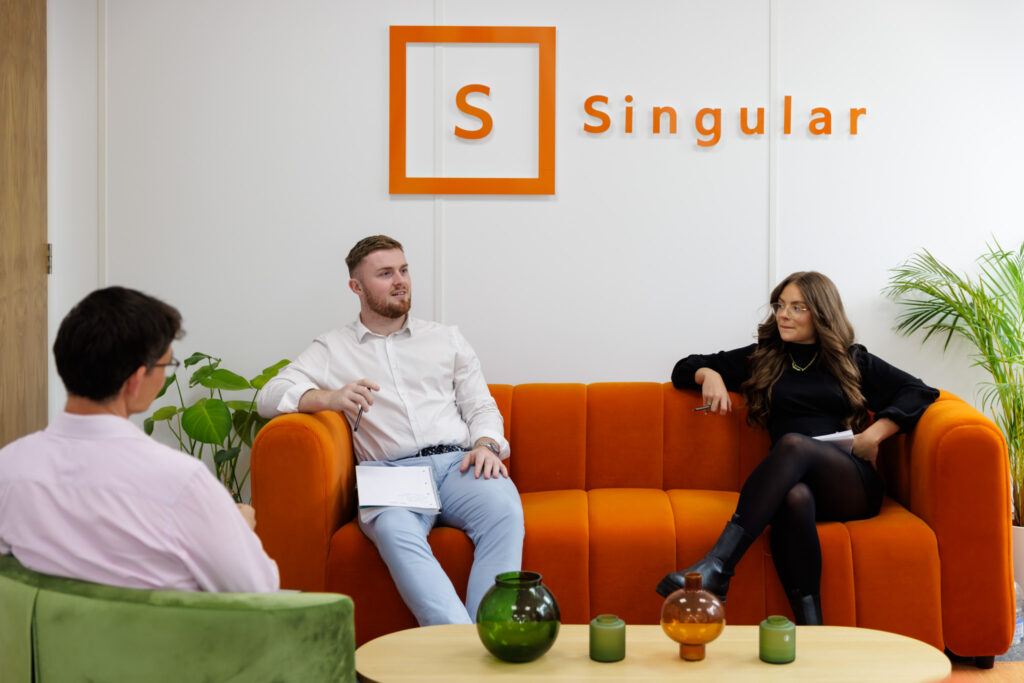 Biotech recruitment: image shows some members of the Singular team sitting on a bright office sofa at the Singular offices, talking to one another. There are green plants either side of the sofa, and the orange Singular logo appears on a white wall behind them.