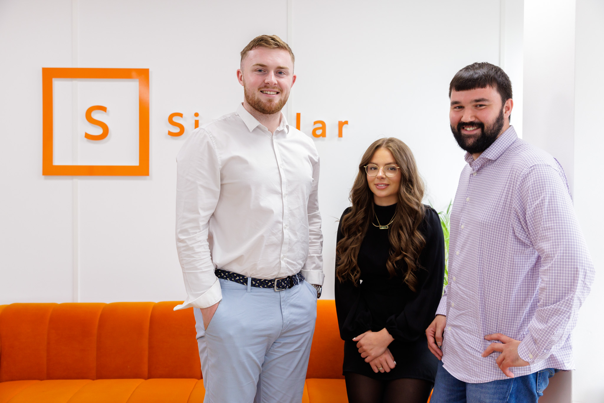 Biotech recruitment: image shows Singular's talent executives, Tyler, Nicole and Isaac, standing against an orange sofa in the Singular offices. The Singular logo can be seen on the wall behind them, also in orange.